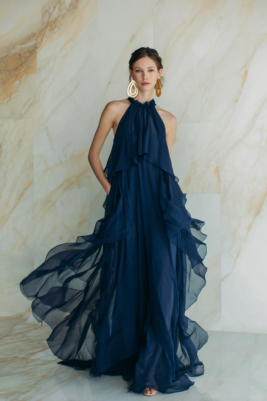 A woman wearing a flowing, floor-length navy blue maxi dress with delicate, layered chiffon fabric, elegant gold hoop earrings, and a sleek updo. The look is completed with minimal makeup and soft natural light illuminating a cream marble backdrop, creating a sophisticated and dreamy atmosphere.