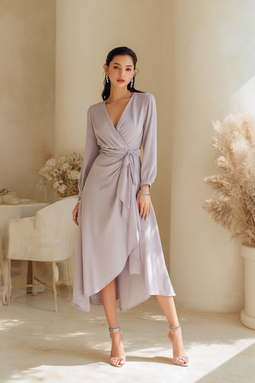 A fashionable woman showcasing a soft lavender wrap dress, cinched at the waist with a matching belt. She pairs this with elegant strappy heels and simple silver bangles. The setting features minimalistic decor with cream tones, illuminated by soft natural light, emphasizing the graceful flow of her dress for an aspirational vibe --ar 2:3 --v 7 --style raw