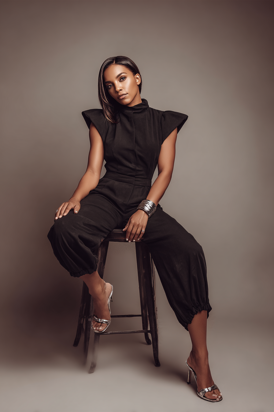 A woman dressed in a sophisticated black jumpsuit with structured shoulders and tailored pants, paired with strappy silver sandals. Her look is enhanced by a statement bracelet and sleek straight hair, posed gracefully in soft studio lighting against a neutral backdrop, representing confidence and elegance.