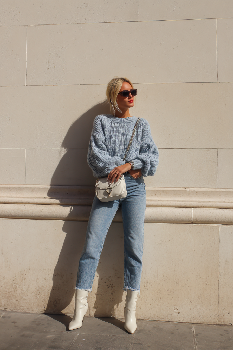 A vibrant look featuring a comfortable oversized sweater in soft blue, tucked into high-waisted denim, completed with stylish ankle boots and a crossbody bag. This casual yet chic outfit is set against a minimalist cream wall, brightened by soft lighting, radiating a relaxed luxury --ar 2:3 --v 7 --style raw