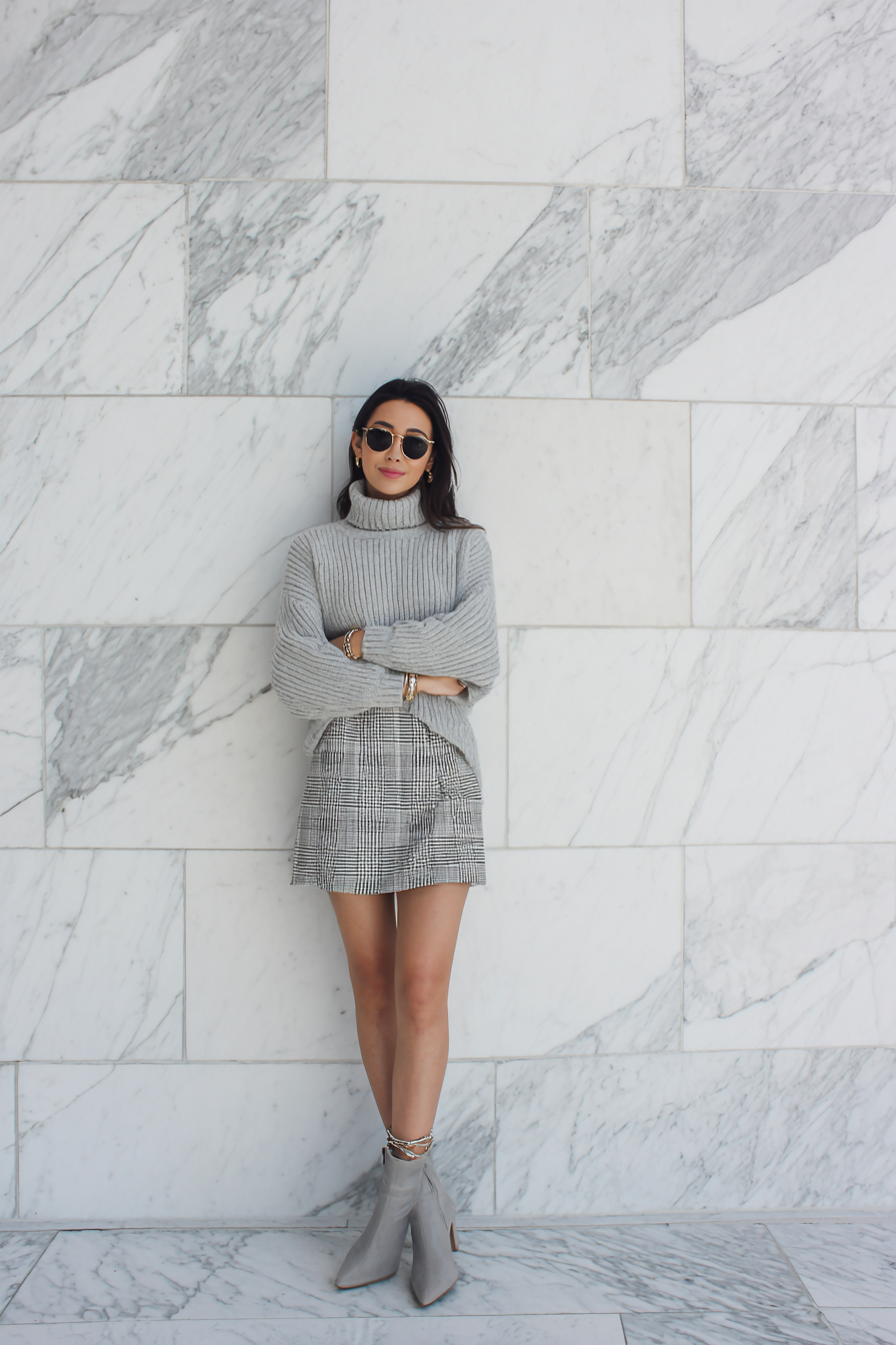 An effortlessly chic outfit featuring a petite woman in a structured turtleneck sweater in soft gray and an A-line plaid skirt, matched with ankle boots and a delicate bracelet. The clean marble background complements the refined color palette, capturing a timeless elegance.